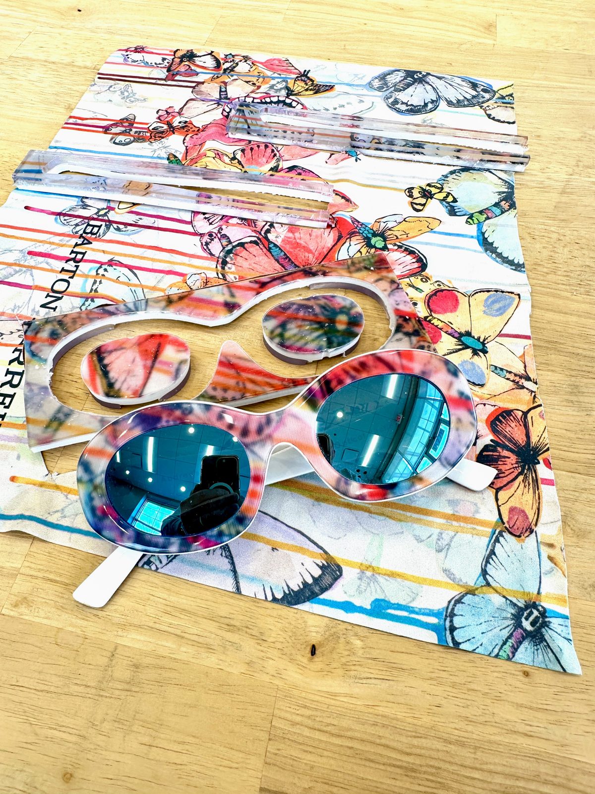Oval bespoke sunglasses with a butterfly print front and white temples, resting on the Barton Perreira cleaning cloth they were built from, with the cut frame front behind them.